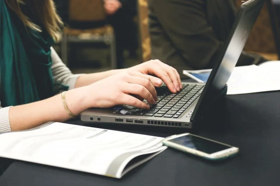 Hands typing on a laptop beside a phone and papers